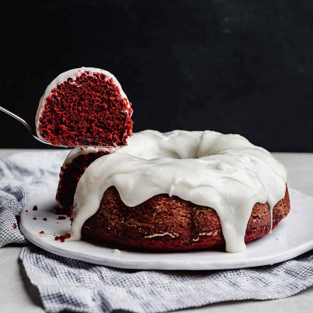 A slice of red valentines day bundt cake hovering over a cream cheese glaze topped Red Velvet Bundt Cake against a black background.