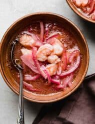 Shrimp Ceviche with sliced purple onions in a ketchup and tabasco based lemon lime sauce, in a bowl on a light gray surface.