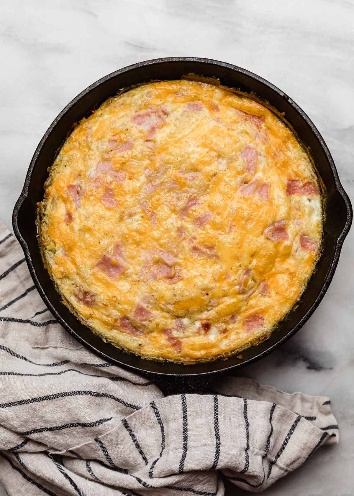 Baked Ham and Cheese Frittata with onion in a round cast iron skillet on a white marble surface.