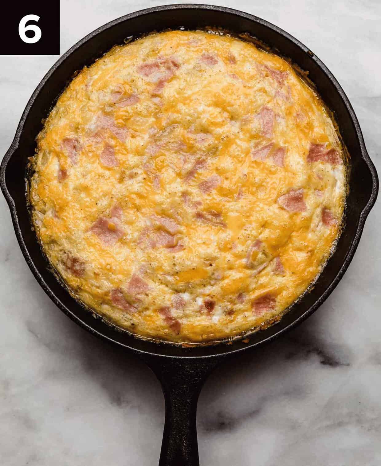 Baked ham and cheese frittata recipe in a cast iron skillet on a white surface.