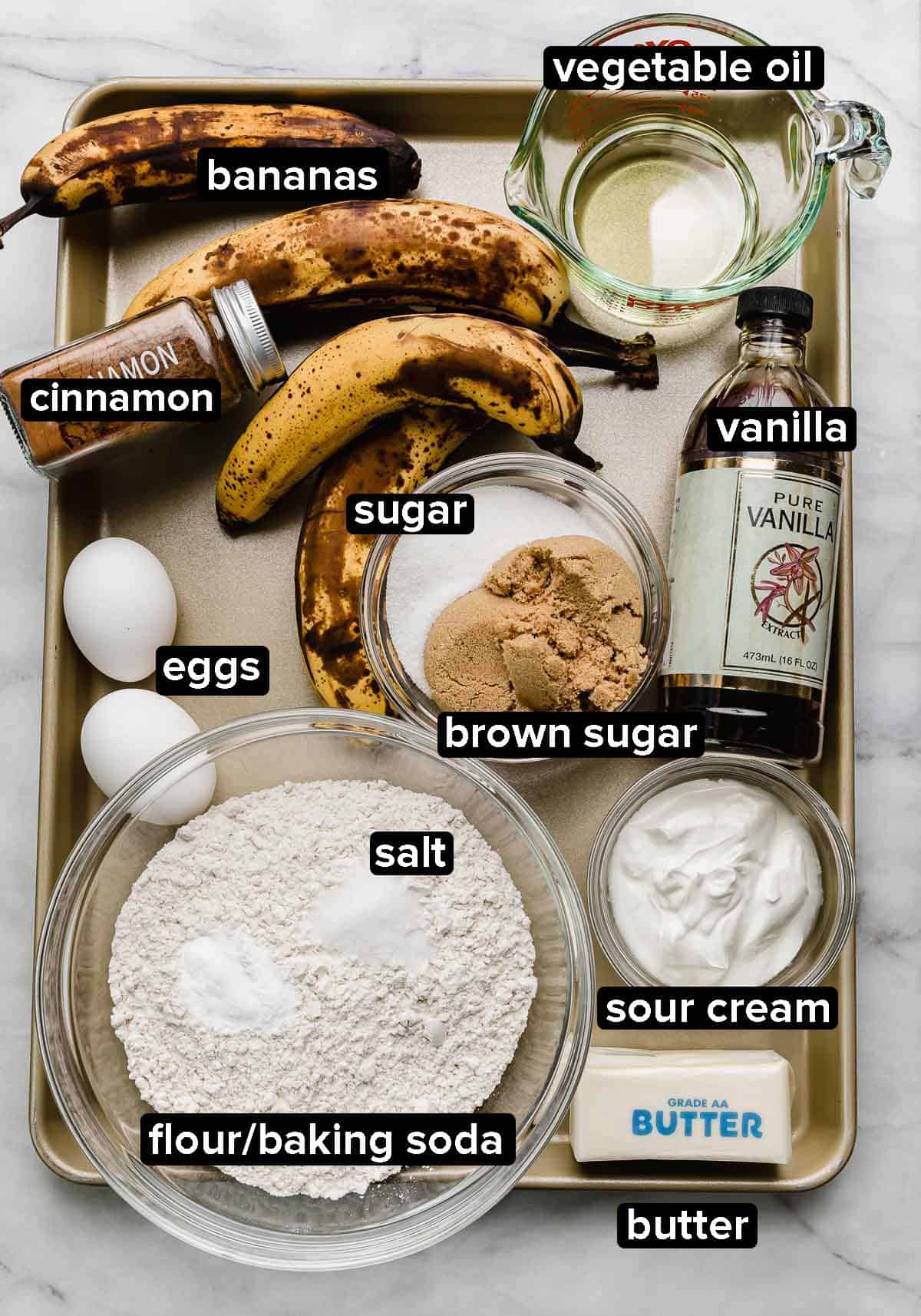 Banana Bread Bundt Cake ingredients on a gold baking sheet: overly ripe bananas, sour cream, butter, eggs, oil, vanilla, sugars, and other dry ingredients.