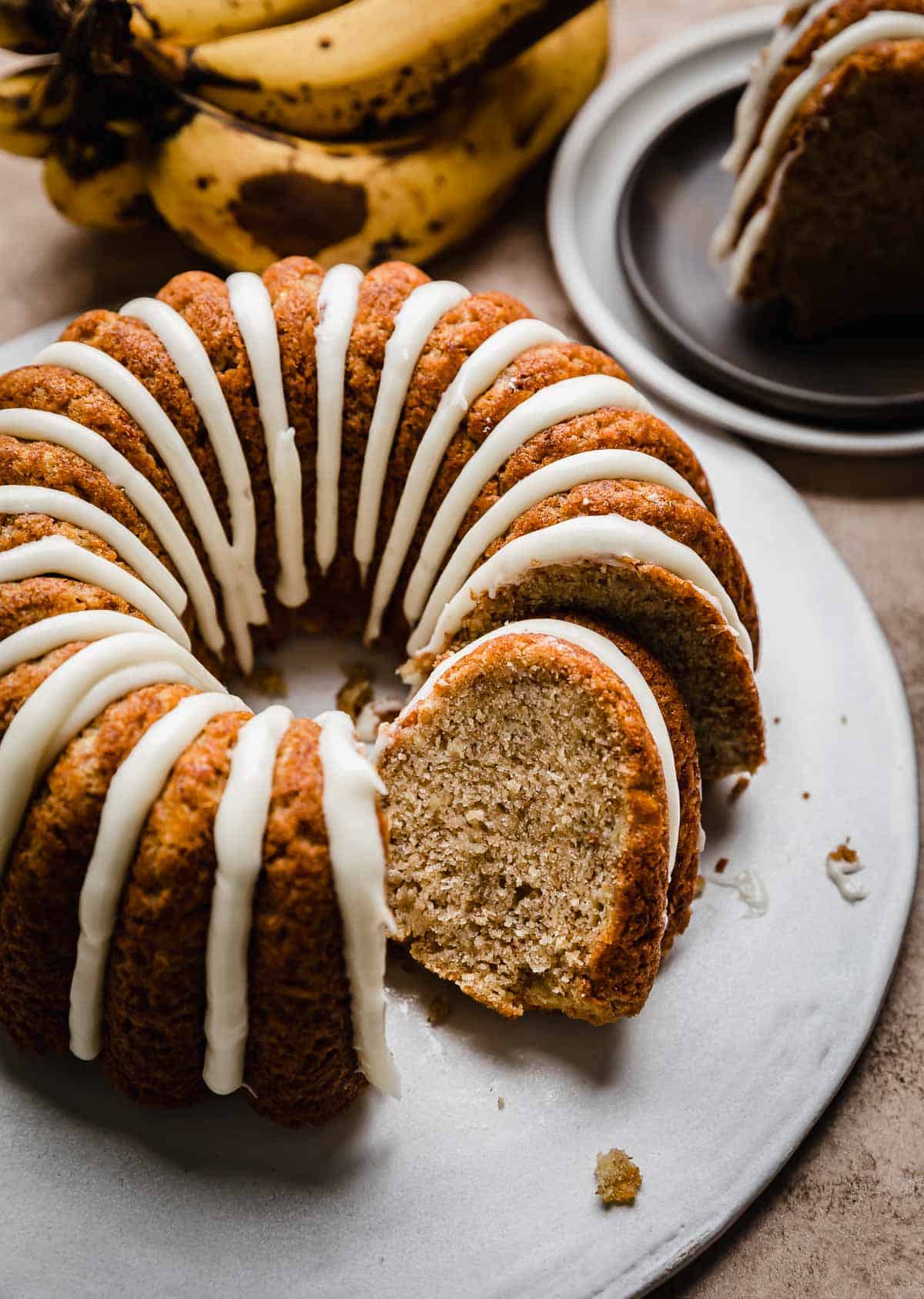 A glazed Banana Bread Bundt Cake on a white cake plate with a slice of banana bundt cake tilting against the full cake.