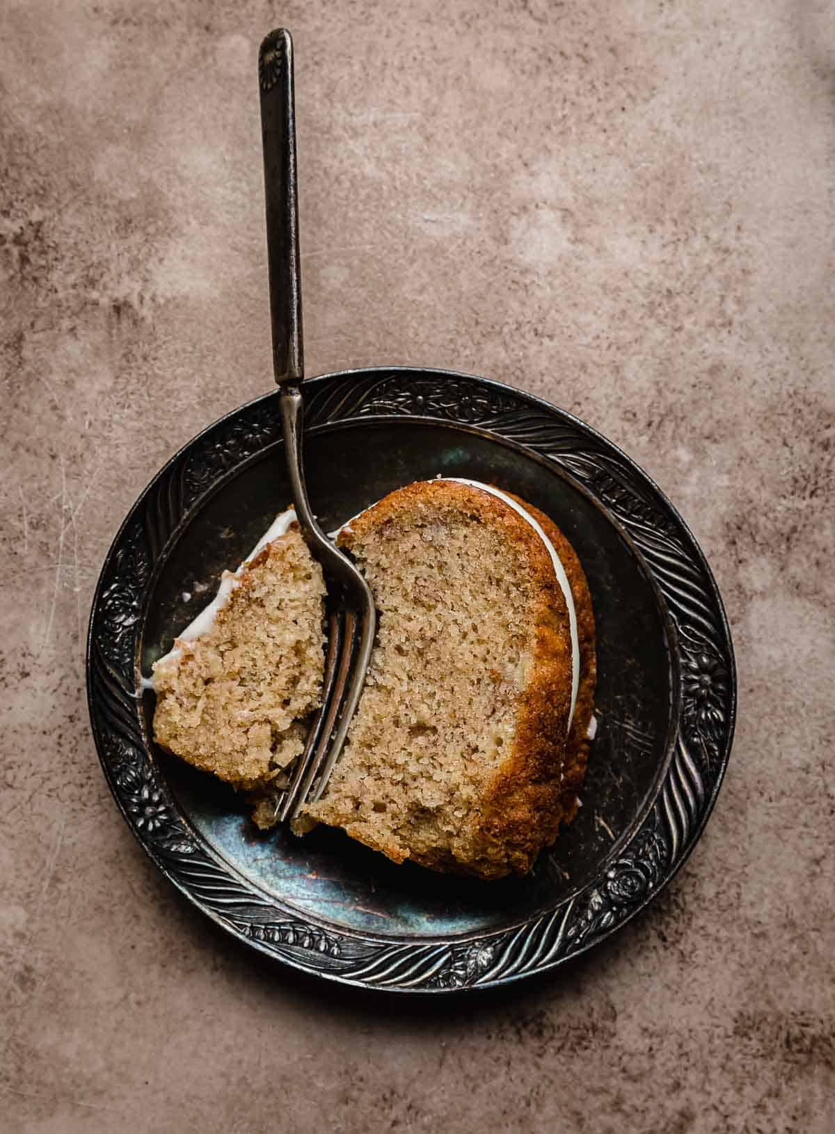 A slice of Banana Bread Bundt Cake on a black antique plate with a fork cutting into the bundt cake.