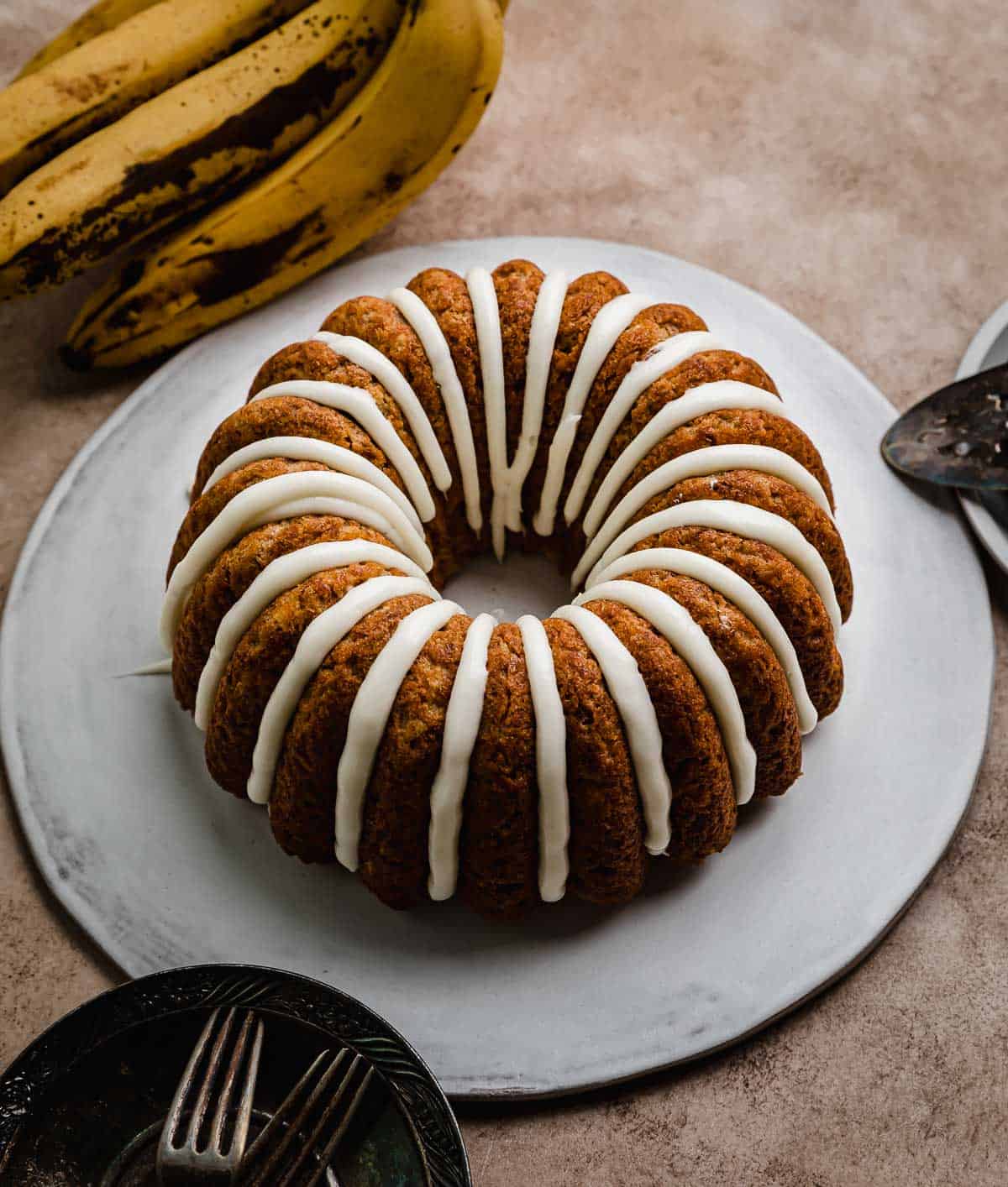 A cream cheese glazed banana bundt cake on a white cake plate that's on a brown textured background.