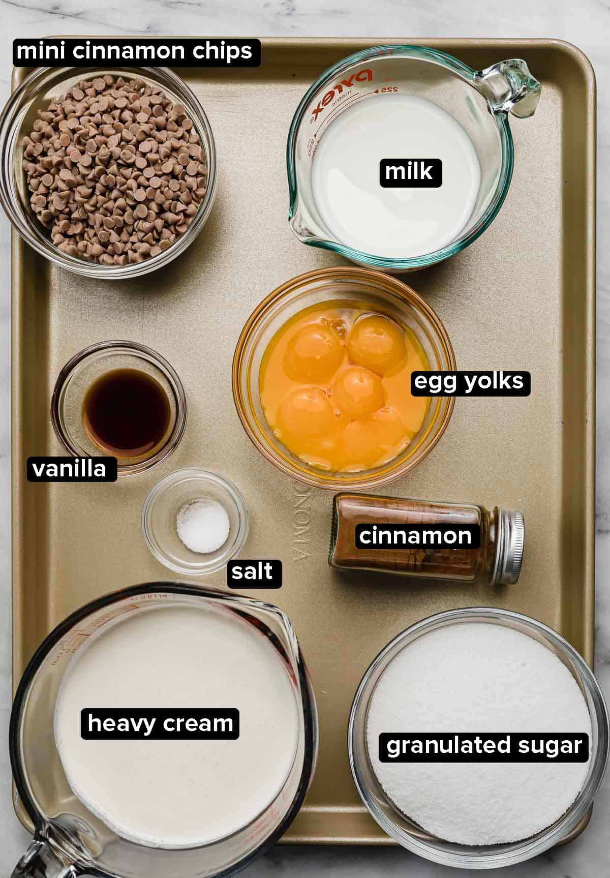Ingredients used to make the best cinnamon spice ice cream on a gold baking sheet: heavy cream, milk, egg yolks, cinnamon spice, salt, vanilla, sugar, and mini cinnamon chips.