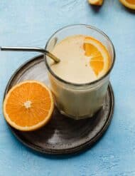 A thick Orange Julius drink recipe in a glass cup with an orange slice in the drink, on a blue background.