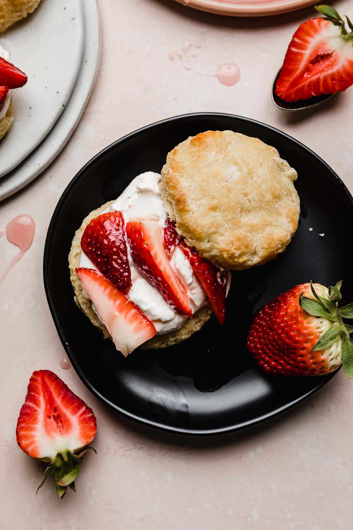 A biscuit topped with whipped cream, sliced strawberries on a black plate, making the best strawberry shortcake recipe ever!
