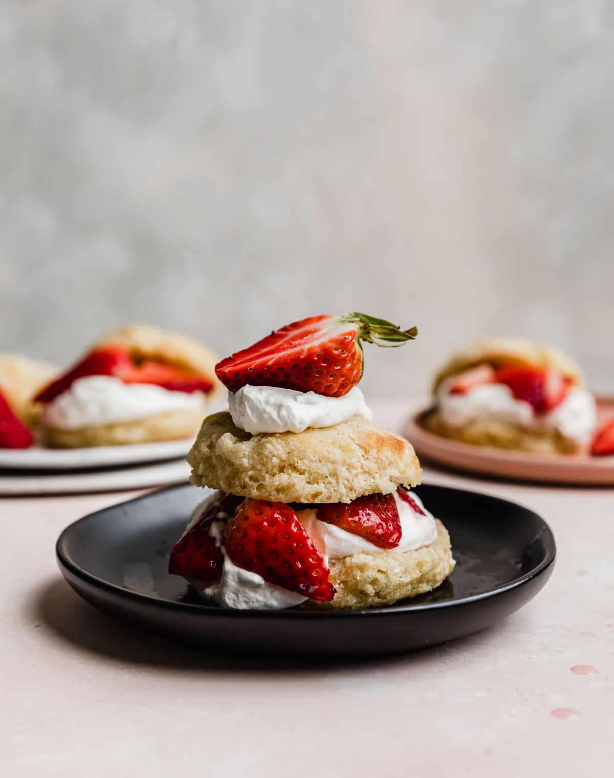A Classic Strawberry Shortcake biscuit cut in half, topped with freshly whipped cream, sugared strawberries, on a black plate.