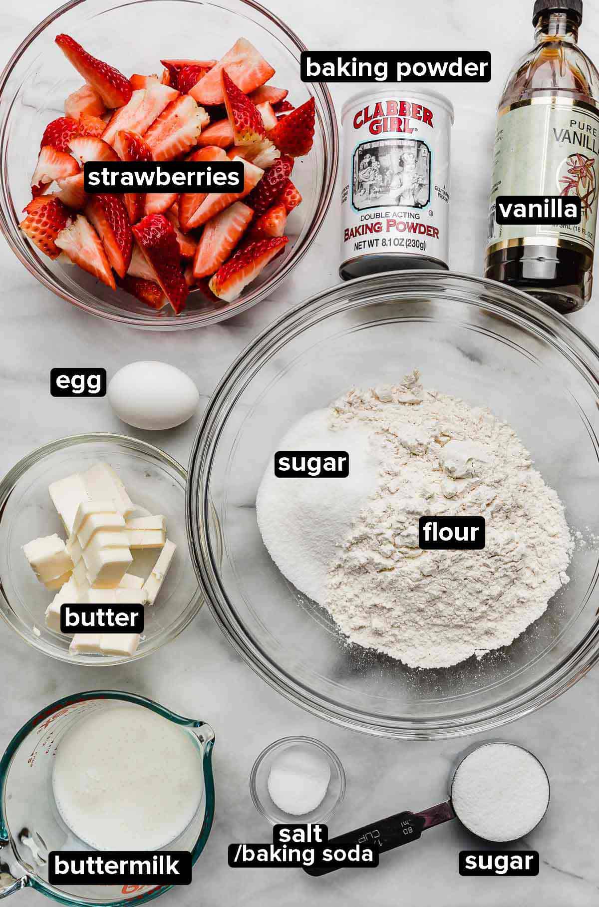 Classic Strawberry Shortcake recipe ingredients portioned into glass bowls on a white marble surface: strawberries, flour, sugar, buttermilk, butter, egg, salt, baking powder, baking soda, and vanilla.