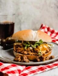 A Cheeseburger Sloppy Joes recipe on a bun with cheese and shredded lettuce with a glass of brown soda in the background.