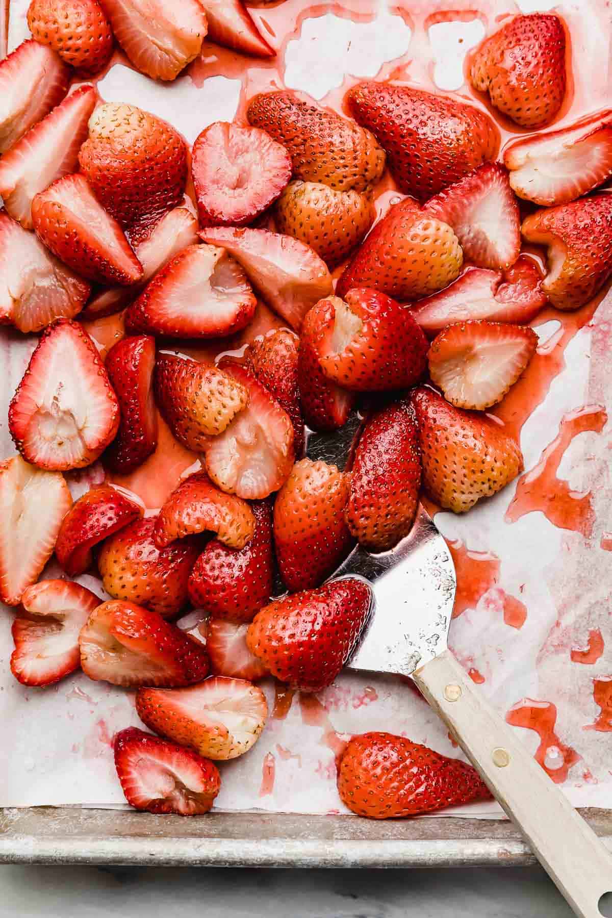 A metal spatula scooping halved baked strawberries from a baking sheet.