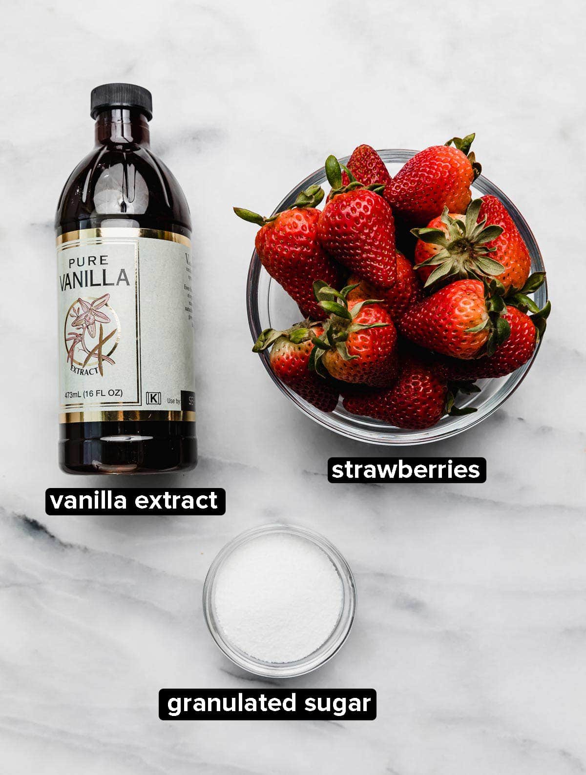 Ingredients used to roast strawberries in the oven: vanilla, fresh strawberries, and granulated sugar on a white surface.