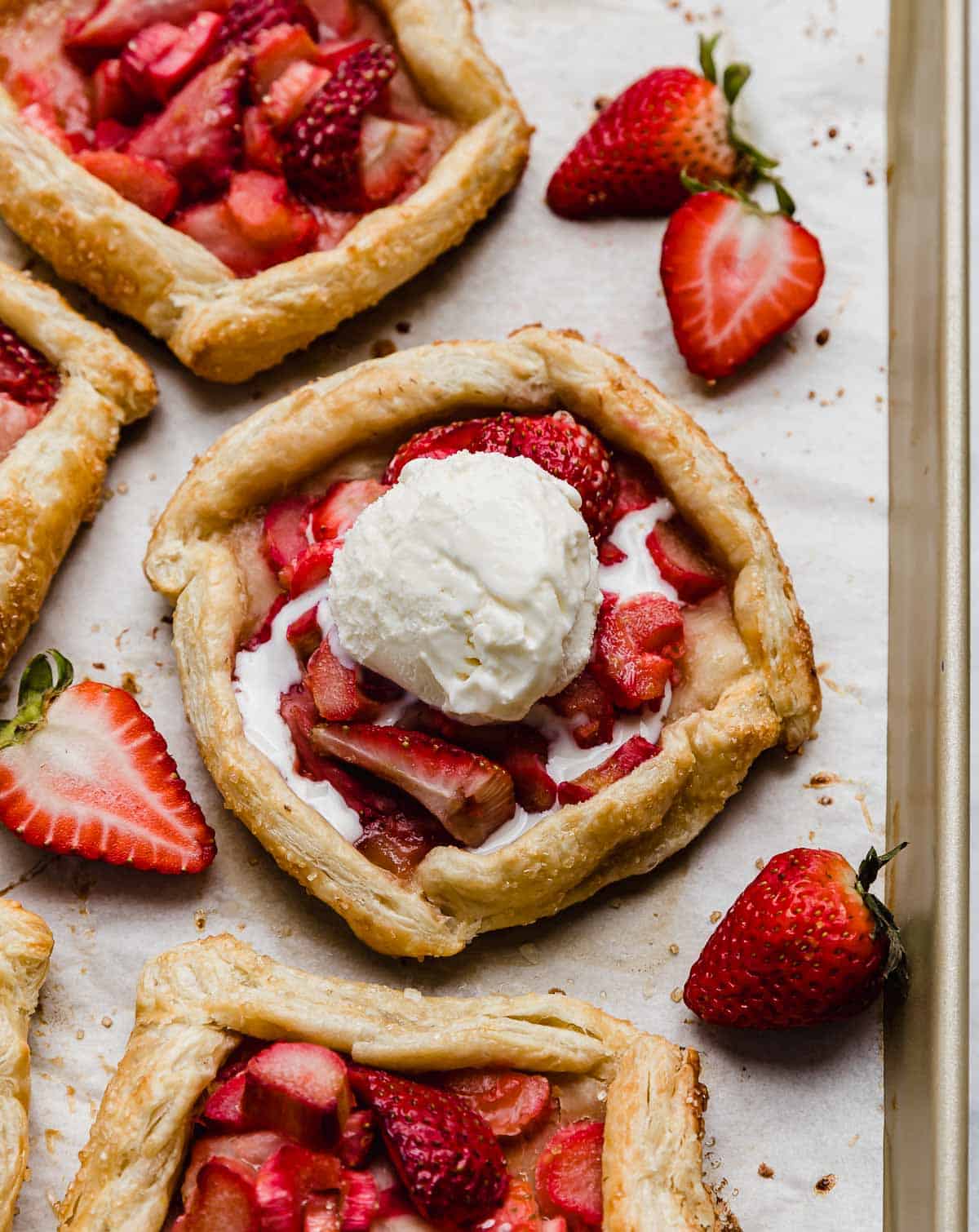 A Rhubarb Strawberry Galette made with puff pastry on a parchment lined baking sheet with a scoop of vanilla ice cream on top of the rustic strawberry rhubarb galette.
