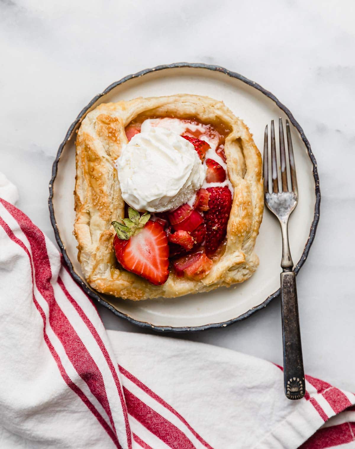 A scoop of vanilla ice cream a top a strawberry and rhubarb galette recipe on a black lined plate on a white marble surface.