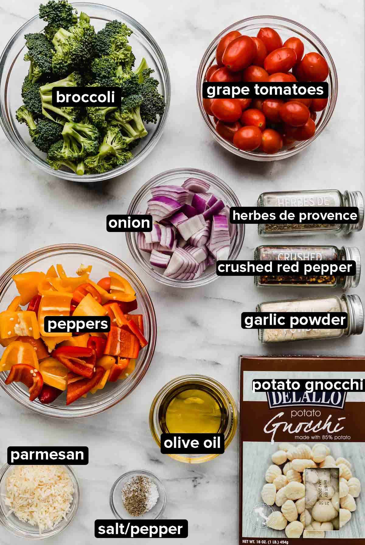 Sheet Pan Gnocchi and Vegetables ingredients portioned into glass bowls on a white marble background: broccoli, peppers, red onion, tomatoes, parmesan, olive oil and gnocchi.