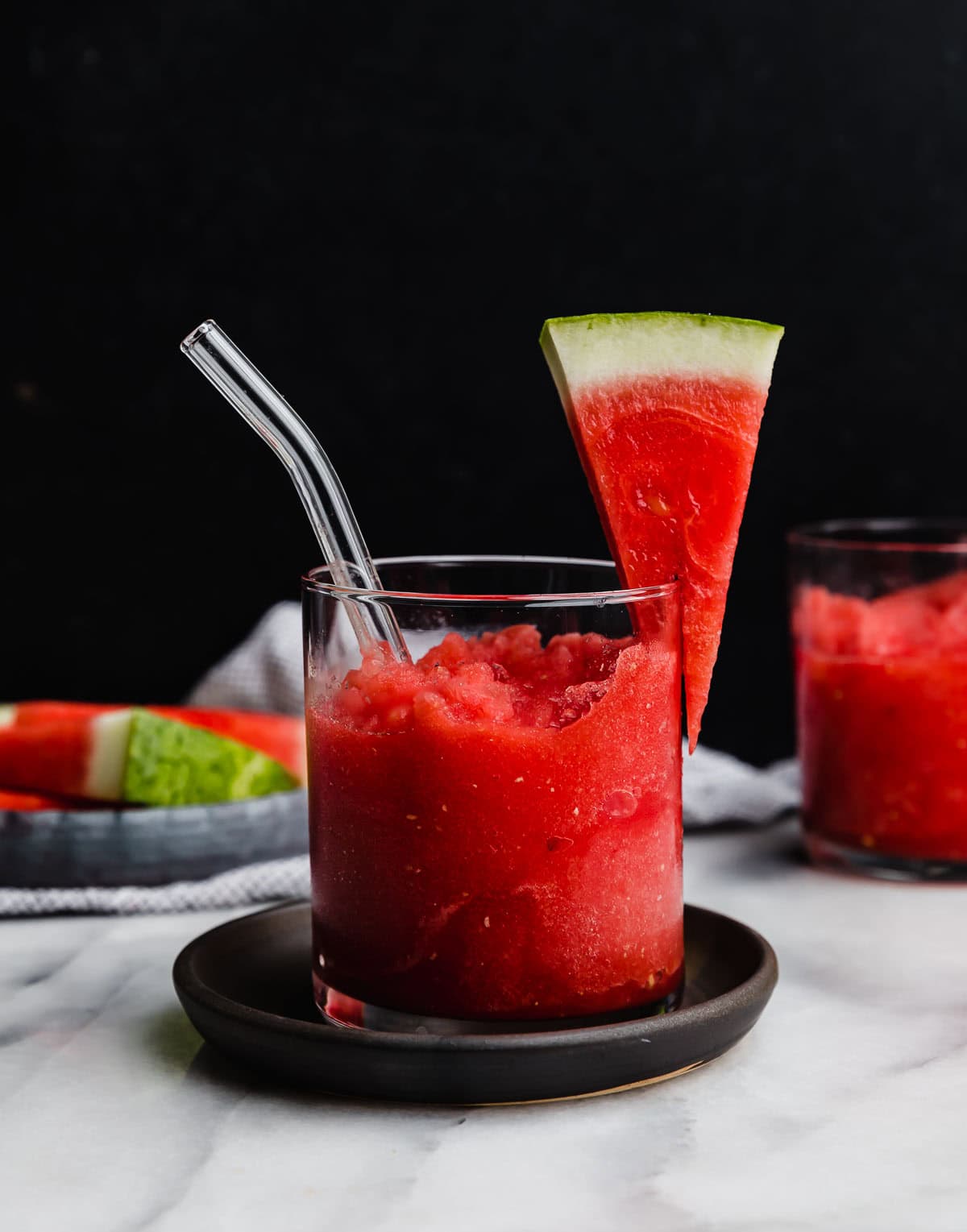 Watermelon Slushie in a glass cup with a glass straw in the cup, against a black background.