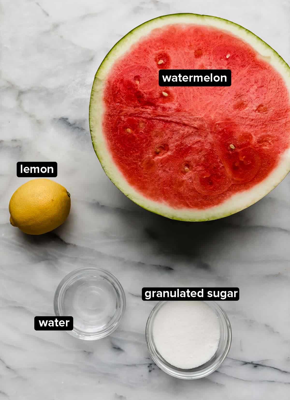 Watermelon Slushie recipe ingredients on a white marble surface: watermelon, lemon, water, and sugar.
