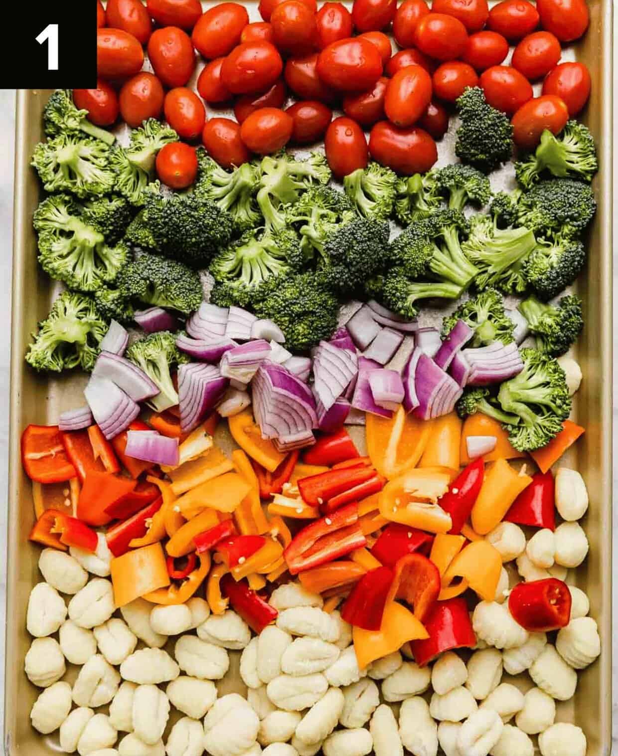 Vegetables and gnocchi on a sheet pan, separated by colors (top to bottom): tomatoes, broccoli, red onion, red and orange peppers, and gnocchi.