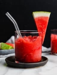 A glass cup filled three quarters full with homemade watermelon slushie with a watermelon wedge on the edge of the cup and a glass straw in the middle of the glass.