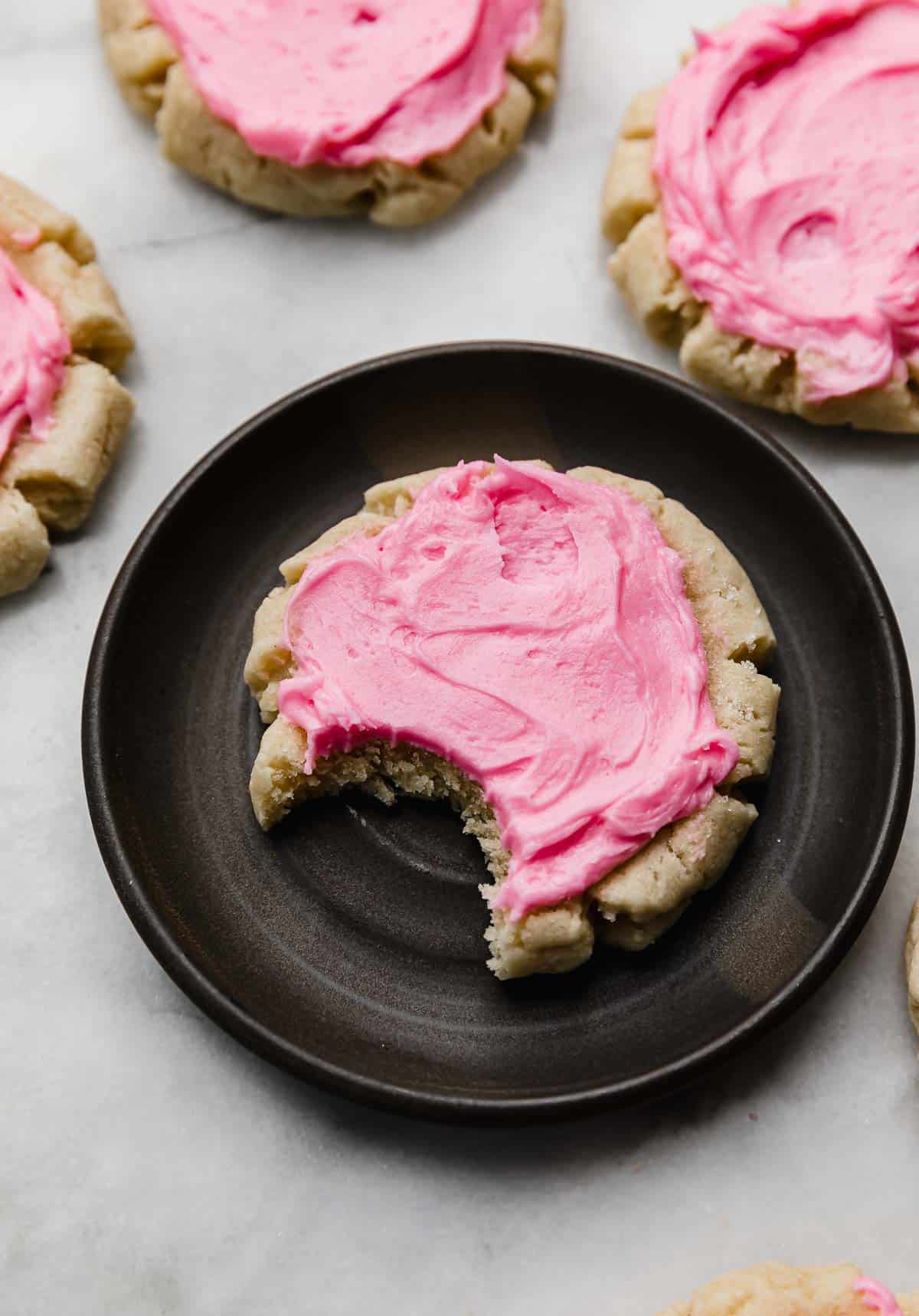A pink frosted Swig sugar cookie with a bite taken out of it on a black plate. 