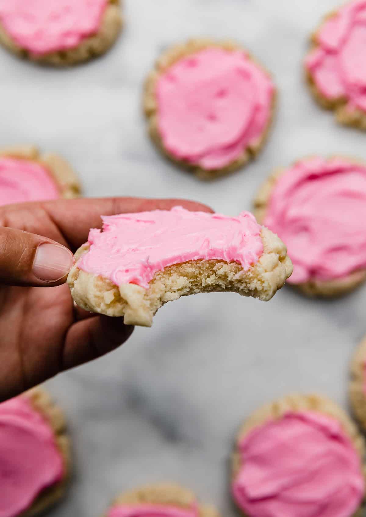 A hand holding a sugar cookie topped with pink frosting, with a bite taken out of it. 