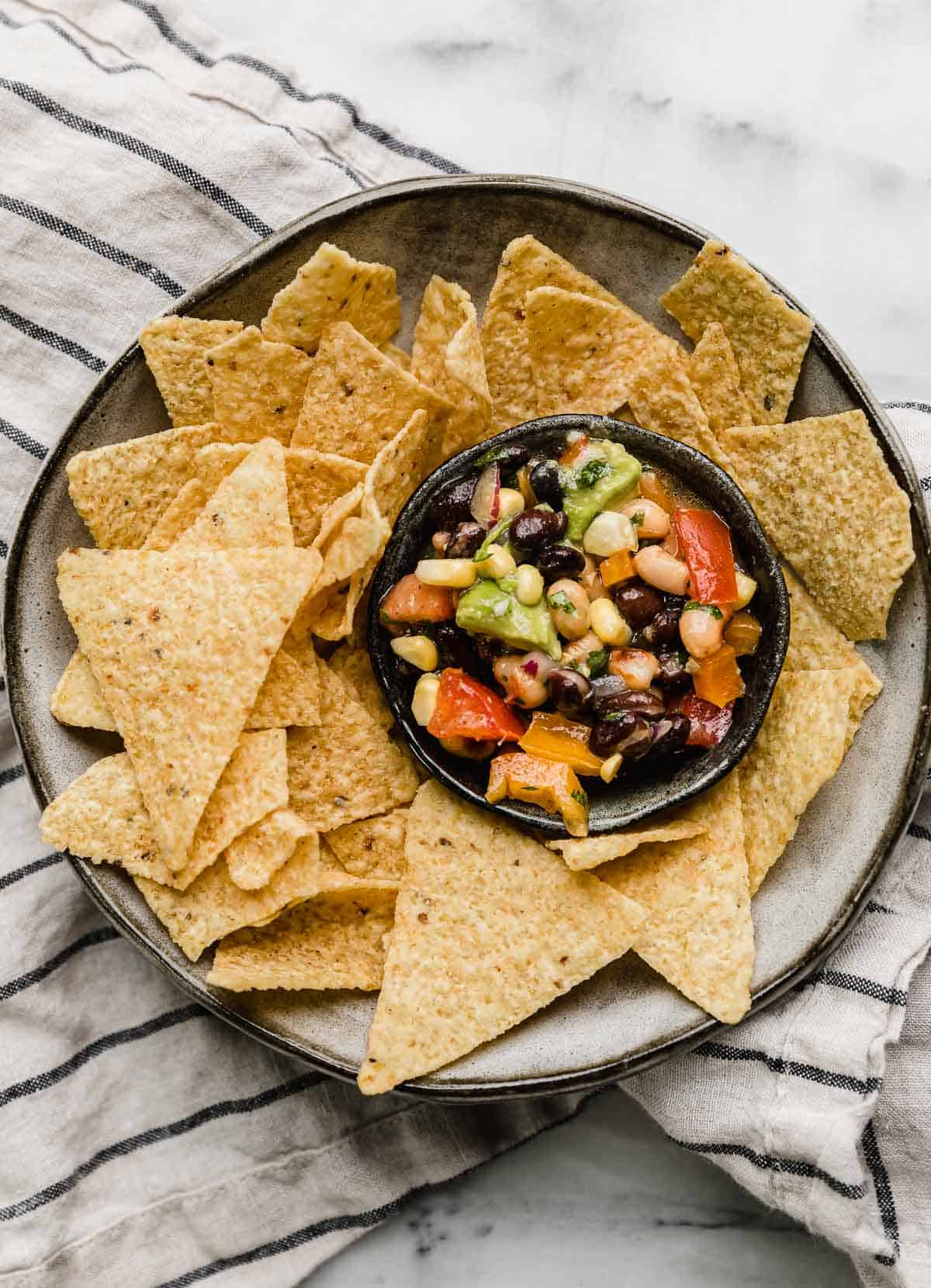 Overhead photo of Texas Caviar in a small black bowl that's on a gray plate surrounded by triangle tortilla chips.