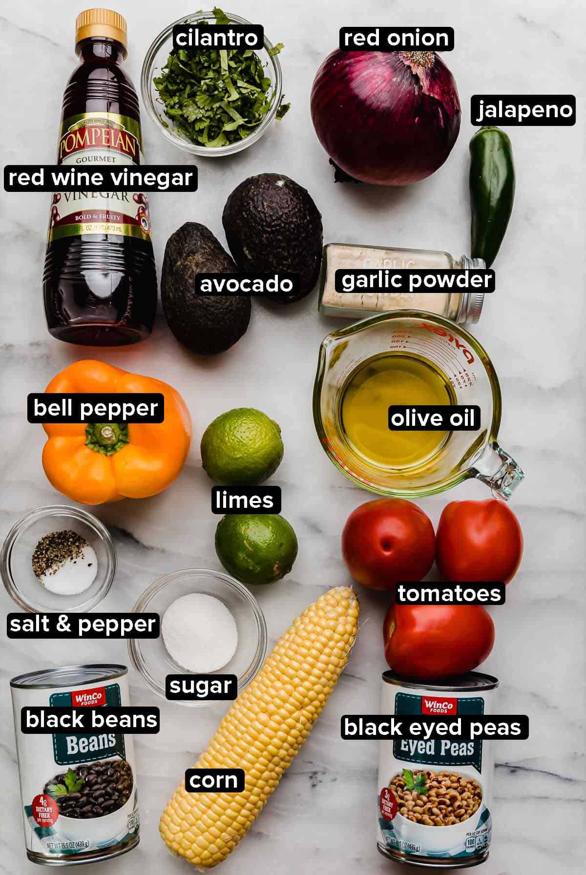 Texas Caviar ingredients on a white marble surface: bell pepper, tomatoes, corn, beans, avocado, cilantro, jalapeño, red wine vinegar, salt and pepper.