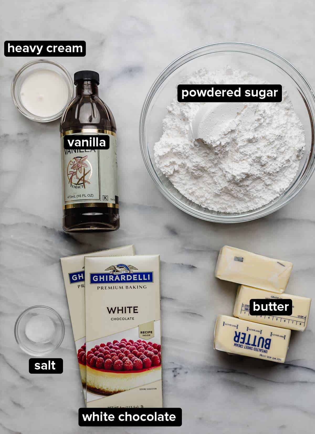 White Chocolate Buttercream Frosting ingredients on a white surface: powdered sugar, white chocolate bars, butter, salt, vanilla, and heavy cream.