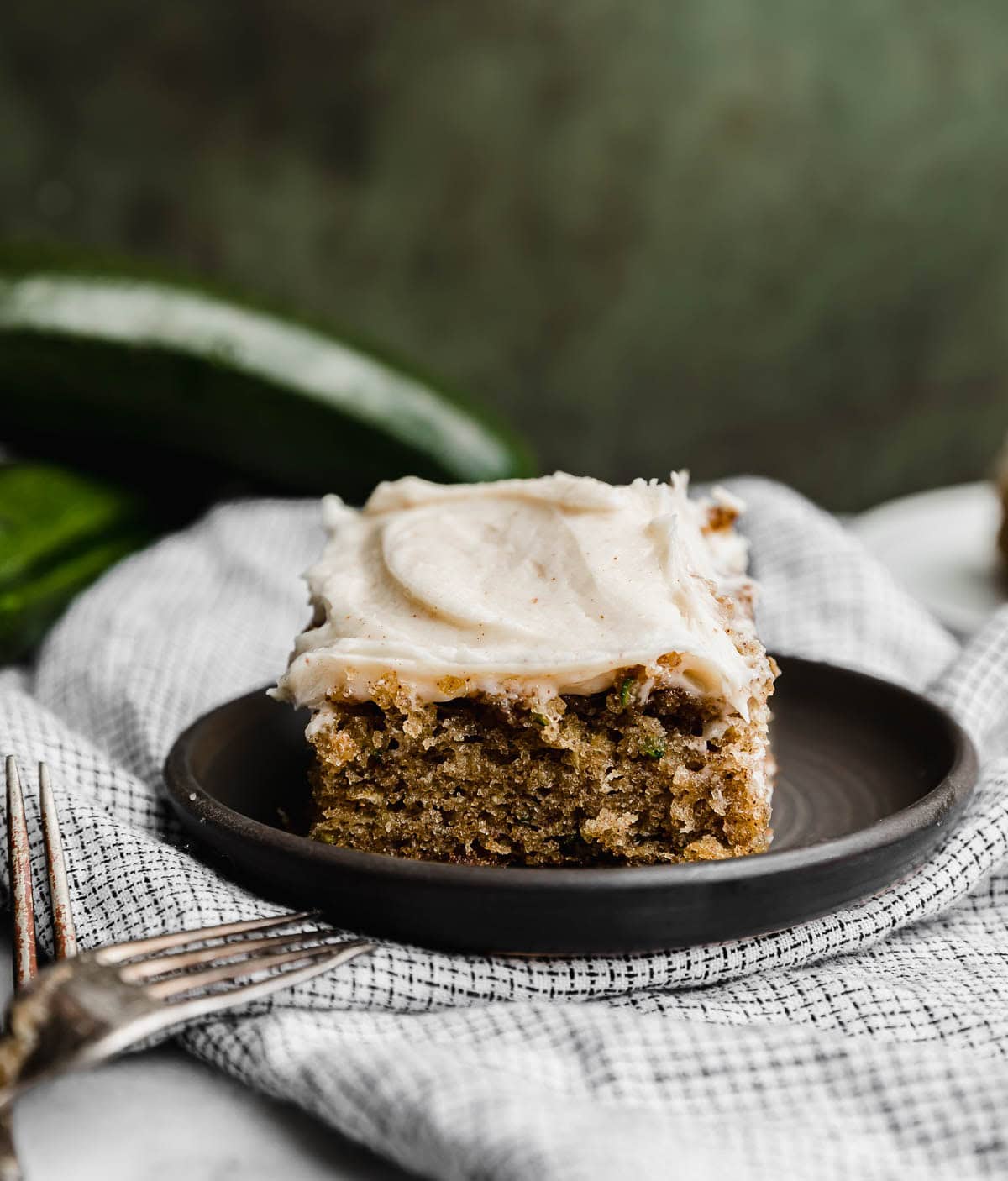 A slice of zucchini cake topped with frosting, on a black plate against a dark green background.