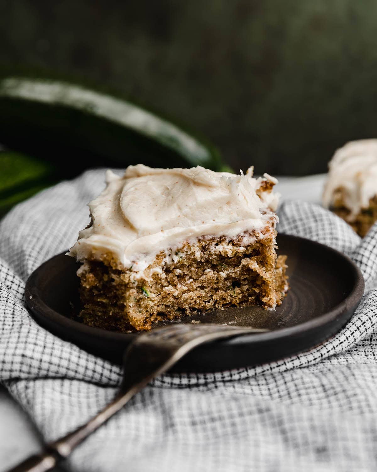A slice of moist zucchini cake on a black plate topped with cream cheese frosting, with a bite taken out of the cake.