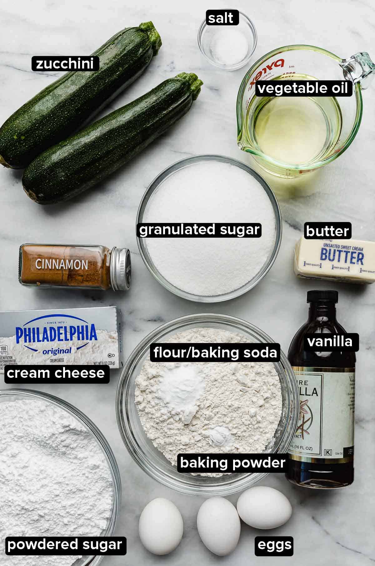 Zucchini cake ingredients on a white surface: shredded zucchini, eggs, vanilla, flour, cinnamon, sugar, butter, salt, oil, and spices.