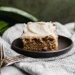 A slice of zucchini cake topped with frosting, on a black plate against a dark green background.