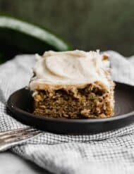 A slice of zucchini cake topped with frosting, on a black plate against a dark green background.