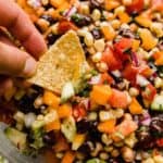 Texas caviar (diced tomatoes, peppers, beans, cilantro, and onion) in a glass bowl with a hand dipping a tortilla chip into the cowboy caviar recipe.