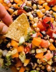 Texas caviar (diced tomatoes, peppers, beans, cilantro, and onion) in a glass bowl with a hand dipping a tortilla chip into the cowboy caviar recipe.