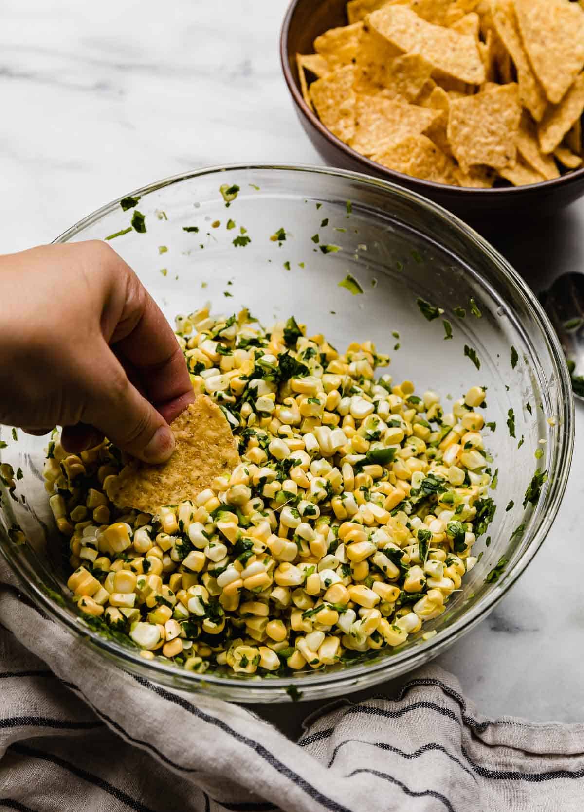 A hand holding a tortilla chip, scooping up some corn salsa with jalapeño and cilantro.