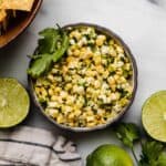 Overhead photo of corn salsa made with raw corn in a black bowl on a white background with fresh limes surrounding the bowl.