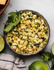Overhead photo of corn salsa made with raw corn in a black bowl on a white background with fresh limes surrounding the bowl.