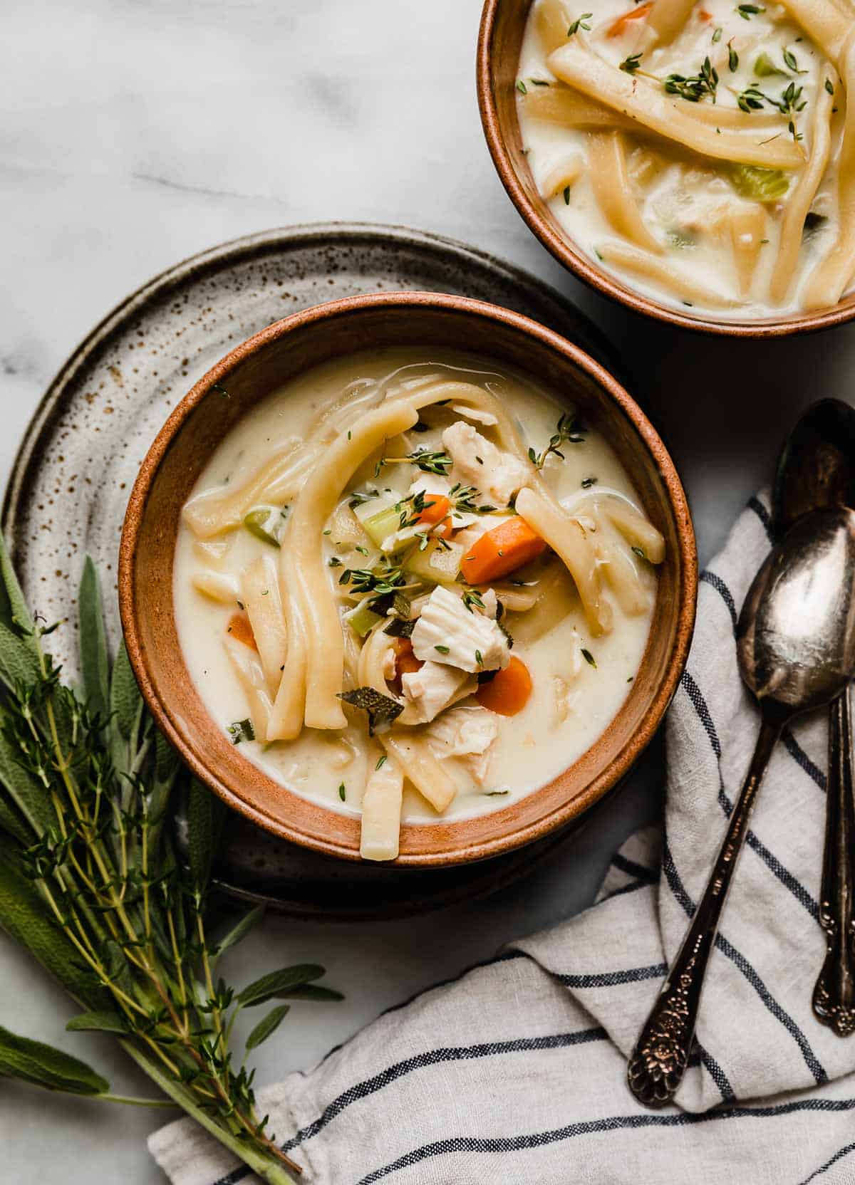Creamy Chicken Noodle Soup in a brown colored bowl on a white marble surface with fresh herbs to the left of the bowl.