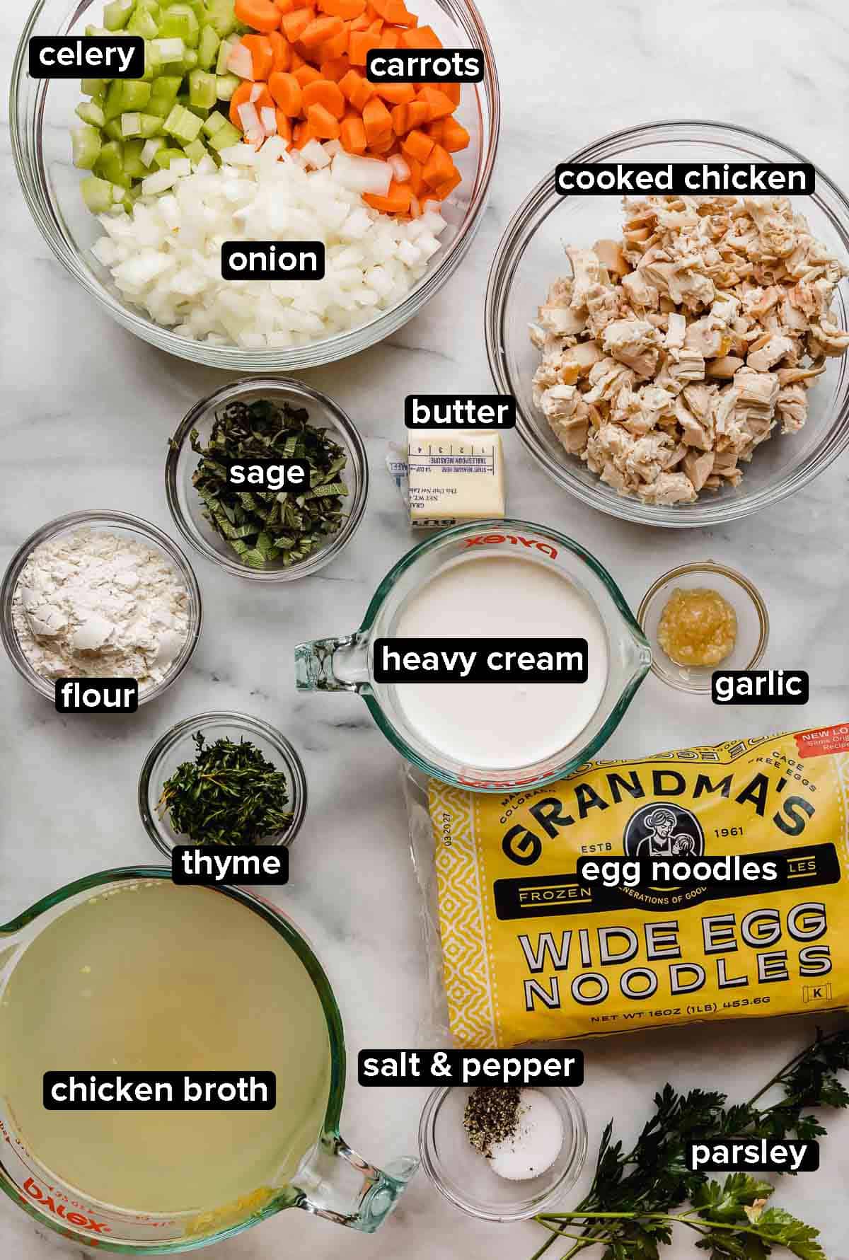 Creamy Chicken Noodle Soup Recipe ingredients on a white marble background: chopped celery and carrots and onions, cooked chicken, sage and thyme and parsley, wide egg noodles, chicken broth, chicken, butter, flour, and heavy cream.