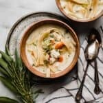 Creamy Chicken Noodle Soup in a brown colored bowl on a white marble surface with fresh herbs to the left of the bowl.