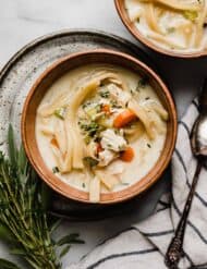 Creamy Chicken Noodle Soup in a brown colored bowl on a white marble surface with fresh herbs to the left of the bowl.