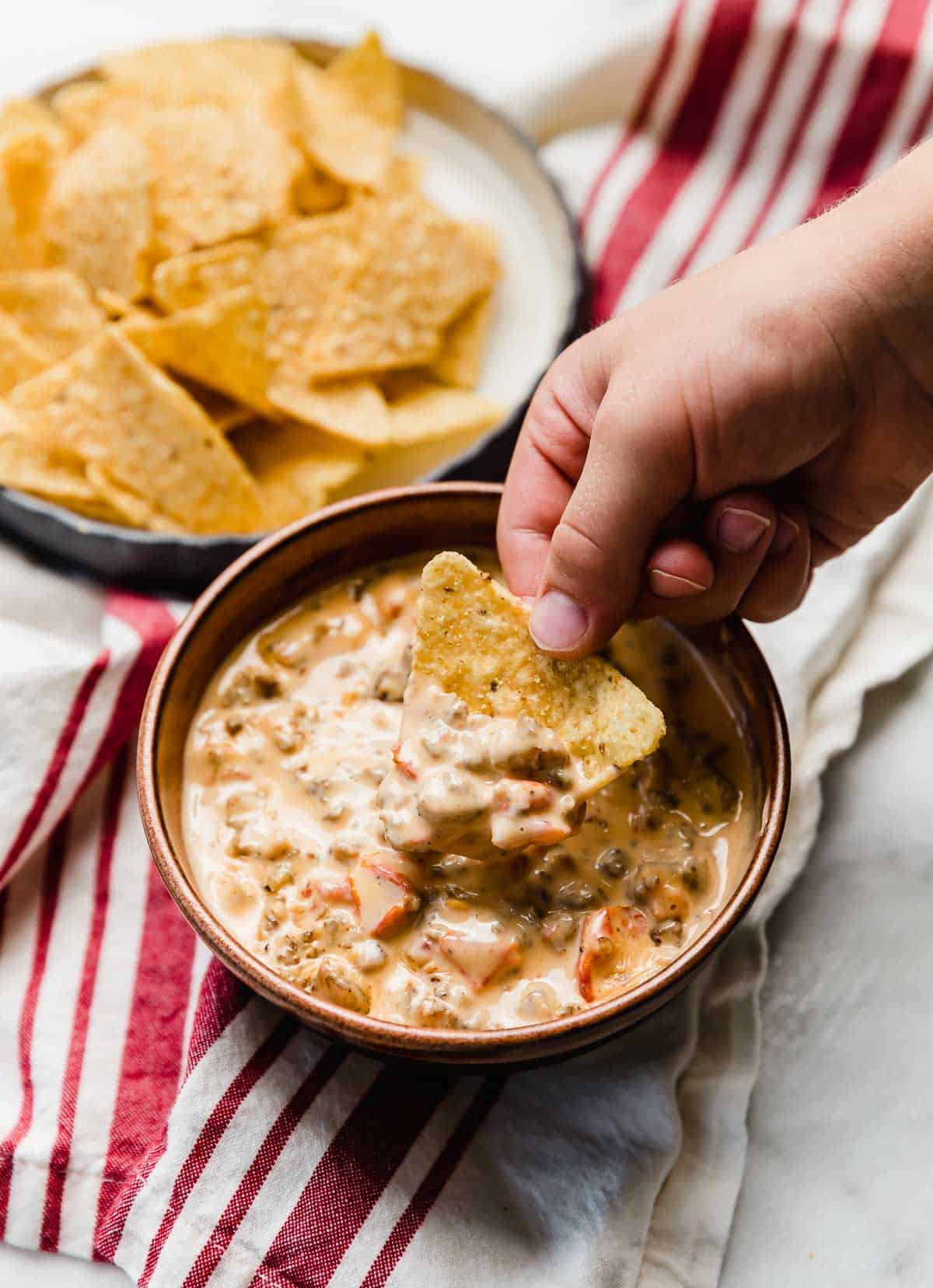 A brown bowl filled with Cream Cheese Sausage Rotel Dip and a hand scooping a tortilla chip into the dip.