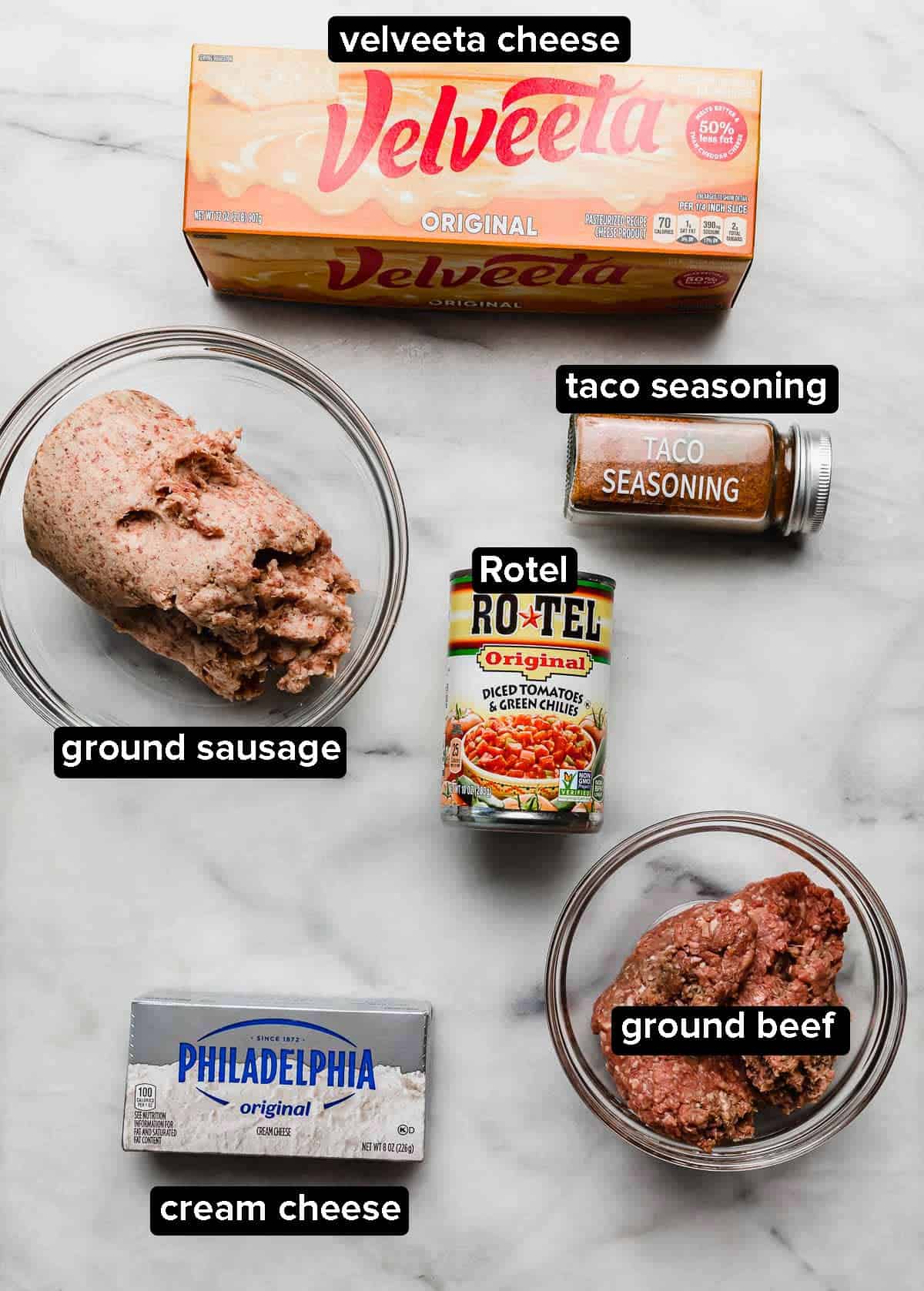 Creamy rotel dip recipe ingredients on a white surface: velveeta cheese, sausage, ground beef, cream cheese, Rotel, and taco seasoning.
